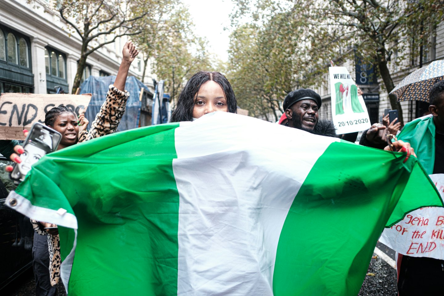 Young woman holds Nigerian flag at outdoor protest with raised fists.